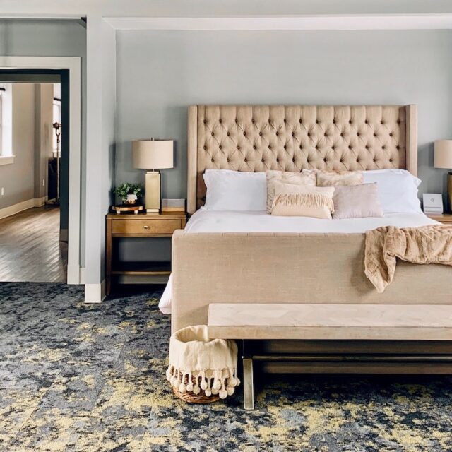 A cozy bedroom in a vacation rental with a comfortable bed with white linens and modern accents, and a soft grey and blue carpet. Gain your competitive edge for your hospitality business with Duchaine Hospitality Services.